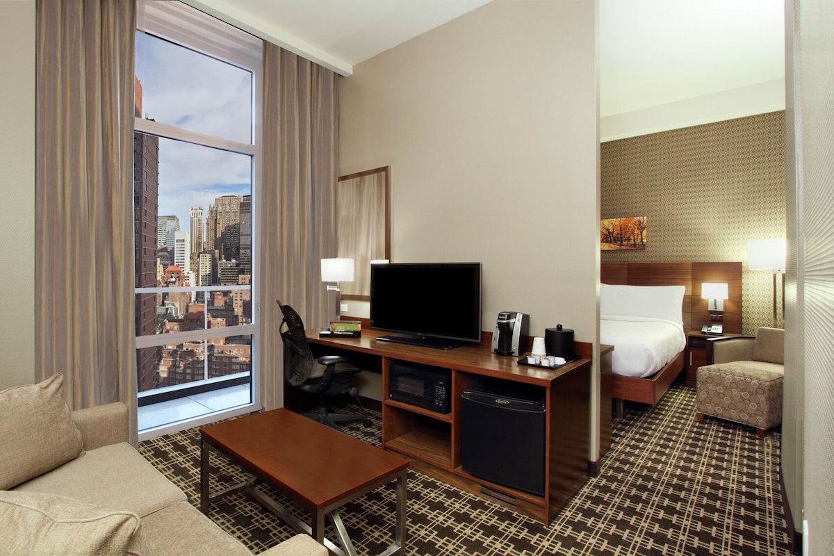 Hotelkamer met groot raam, flatscreen televisie, bureau met koffiezetapparaat, magnetron en bed met hoofdbord.