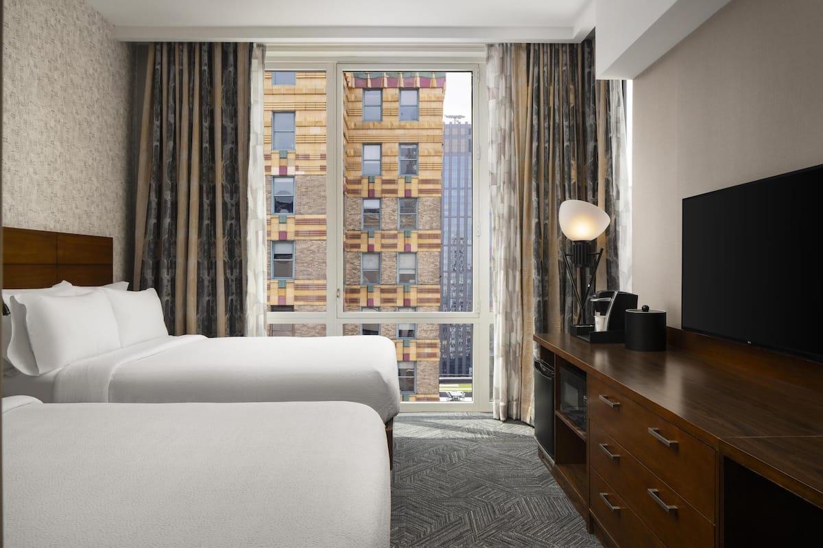 Habitación de hotel con dos camas, un ventanal grande con cortinas, un televisor de pantalla plana, un aparador de madera y una lámpara.