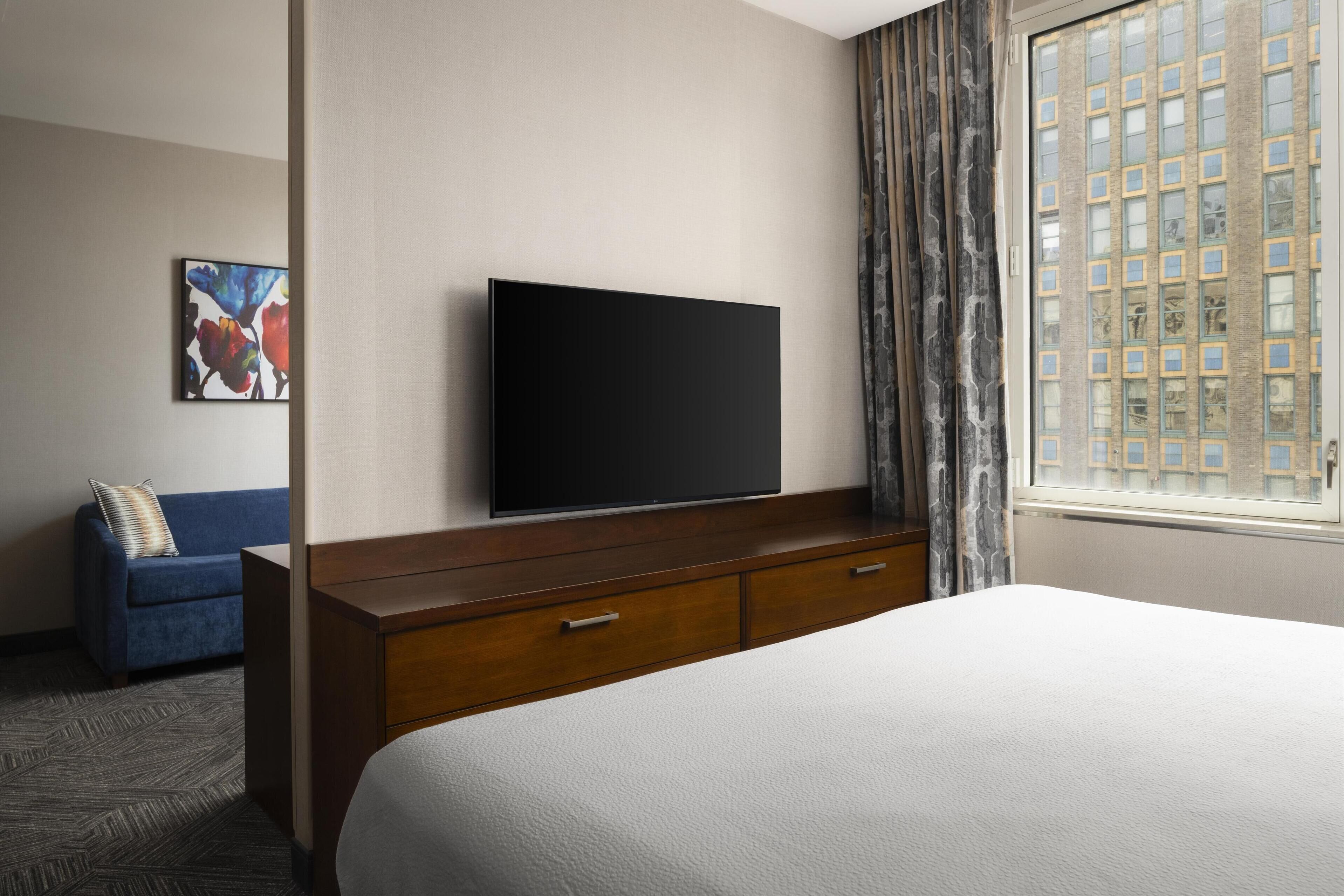 A hotel room with a large bed, a flat-screen TV mounted on a wooden dresser, a blue sofa with a pillow, and a window with a view of buildings.
