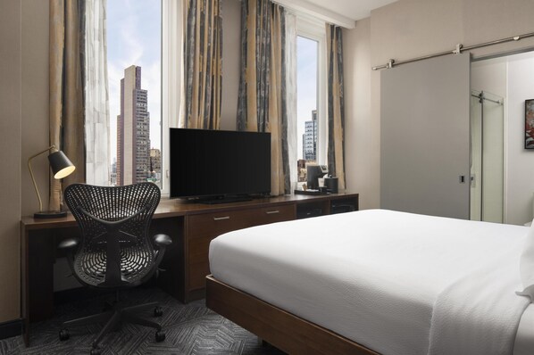 Habitación de hotel con cama, escritorio, silla, televisor y vistas a la ciudad a través de amplios ventanales.