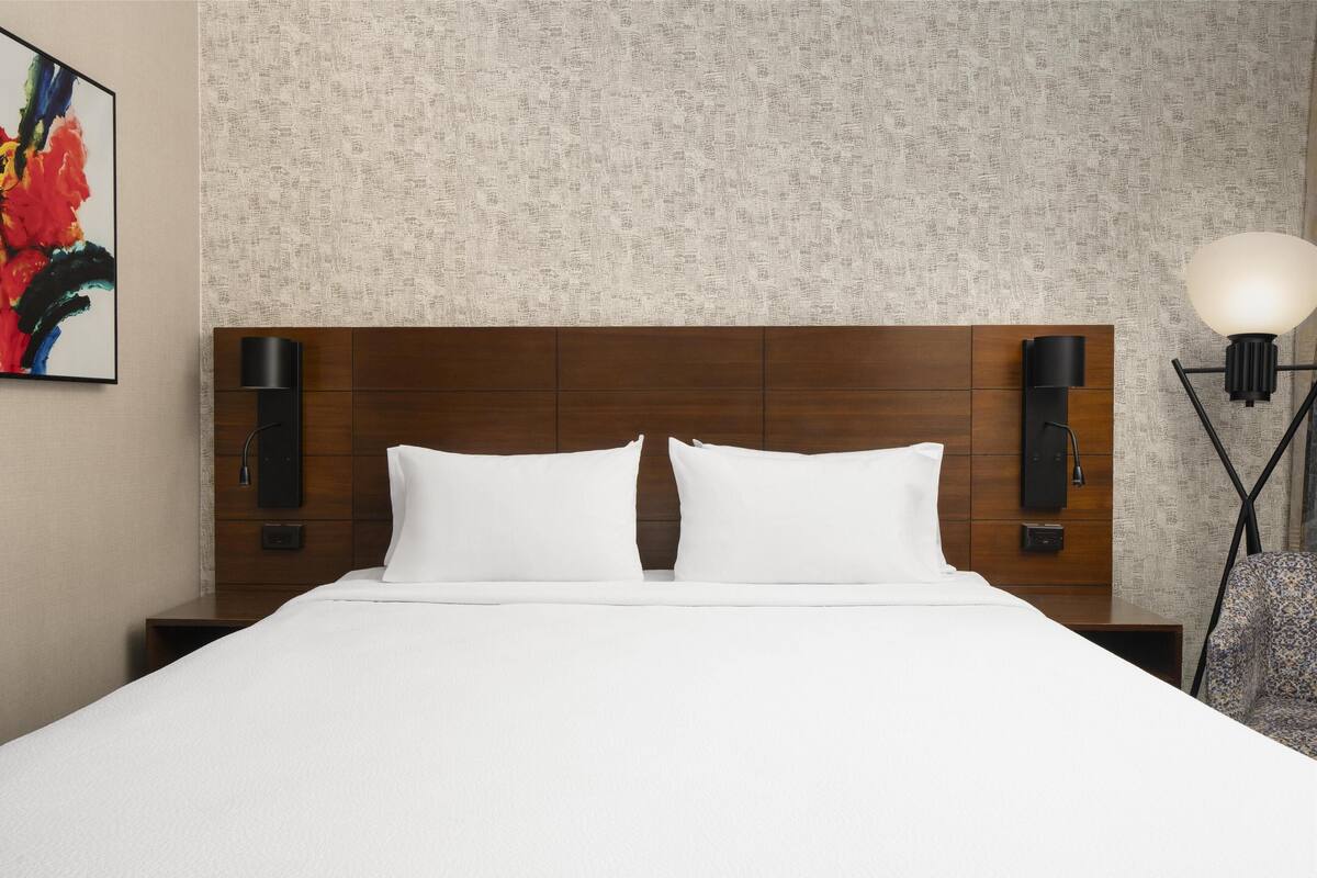 Habitación de hotel con cabecera de madera, ropa de cama blanca, lámpara de pared y una obra de arte enmarcada.