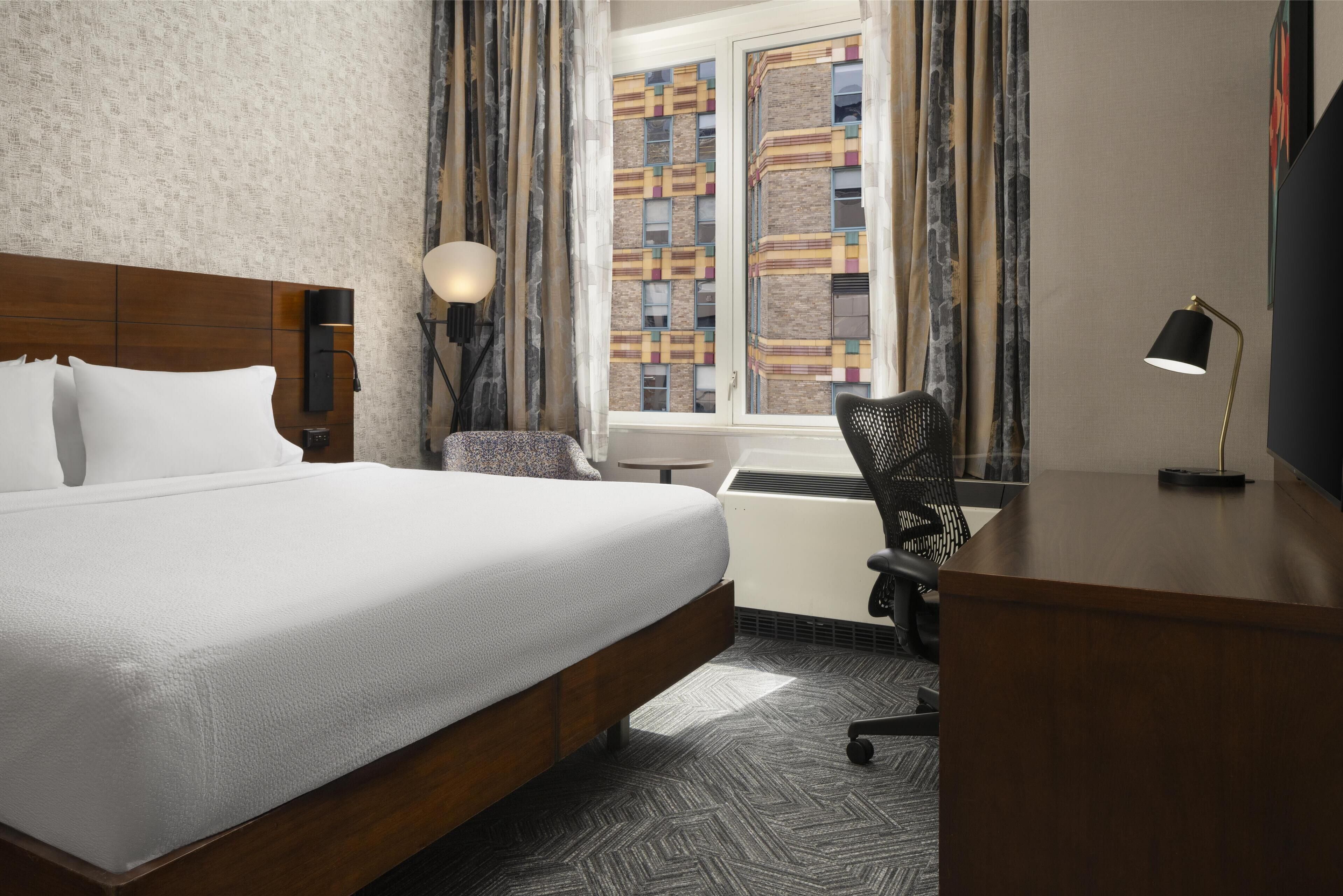 Habitación de hotel con una cama grande, un escritorio, una silla, una lámpara y un ventanal con cortinas estampadas.