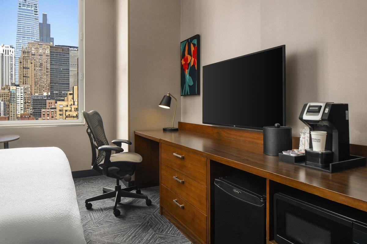 Habitación de hotel con un ventanal grande, un escritorio con computadora, una silla, máquina de café y televisor de pantalla plana.