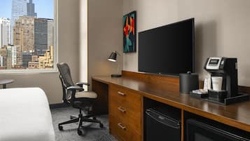 Habitación de hotel con un ventanal grande, un escritorio con computadora, una silla, máquina de café y televisor de pantalla plana.