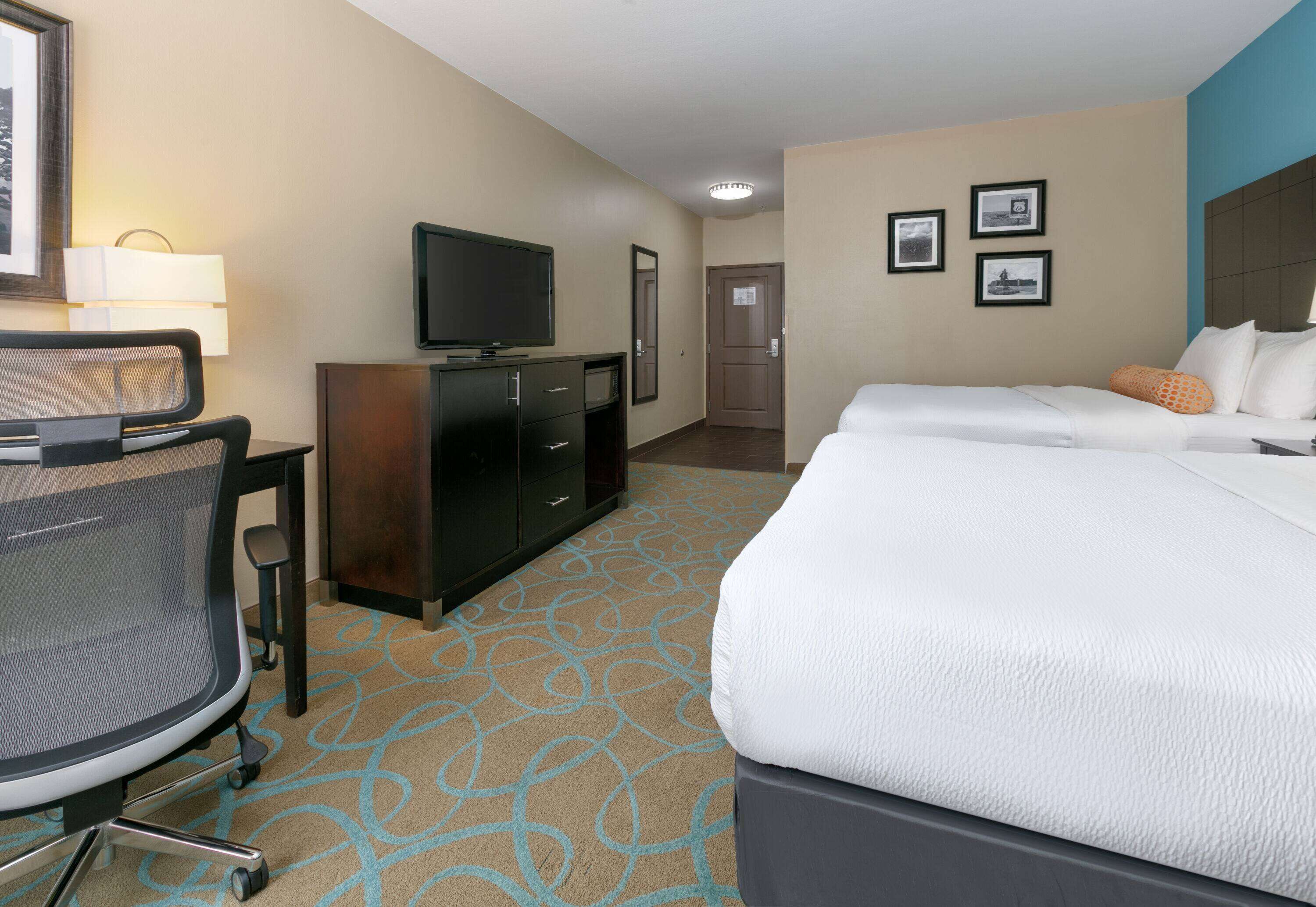 A hotel room with a bed, a desk with a chair, a television, a dresser, and framed pictures on the wall.