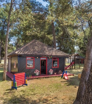 A small wooden building with a red door and windows, surrounded by trees and a playground with a slide. - Apartments Katoro Plava Laguna (Umag)