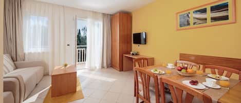 A modern hotel room with a sofa, dining table, breakfast setup, and a TV on the wall.