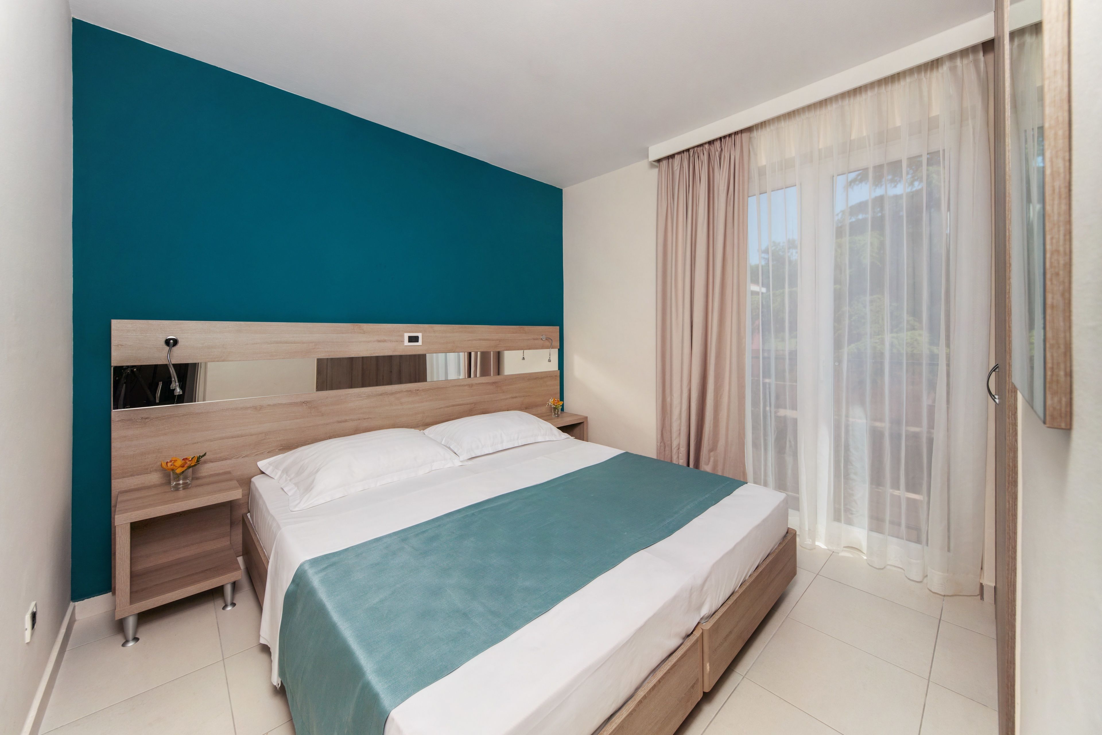 A modern bedroom with a teal accent wall, a wooden headboard, a bed with white linens, and a nightstand with a vase of flowers.