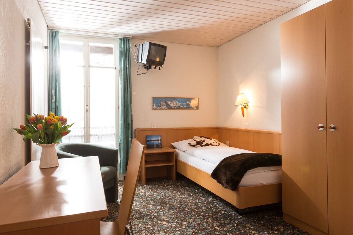 Une petite pièce agréable, dotée d’un lit, d’un bureau, d’une chaise, d’un téléviseur fixé au mur et d’une fenêtre avec des rideaux.