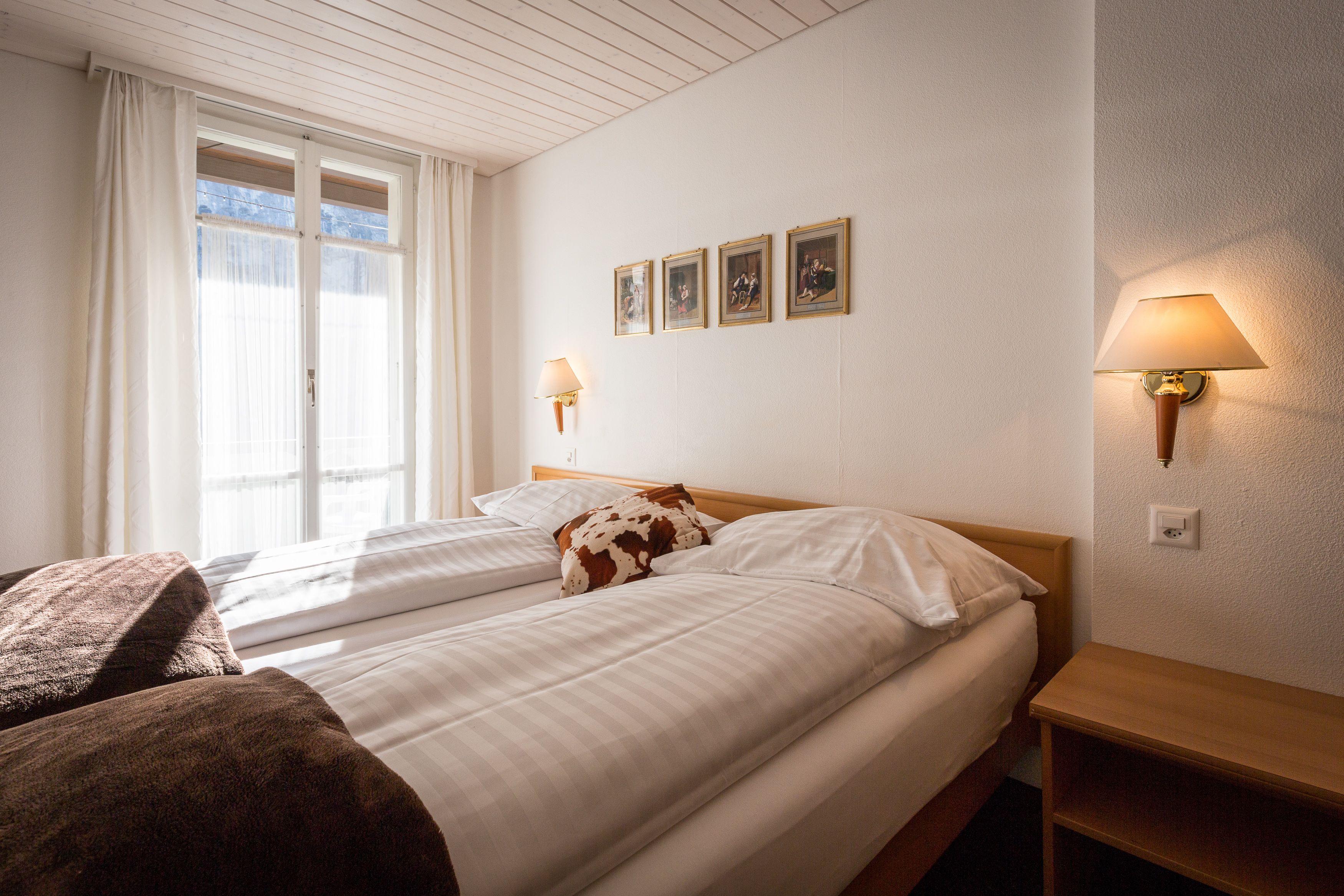 Ein Hotelzimmer mit zwei Betten, einem hölzernen Kopfteil, einem Nachttisch, einer Lampe und einem Fenster mit Vorhängen.