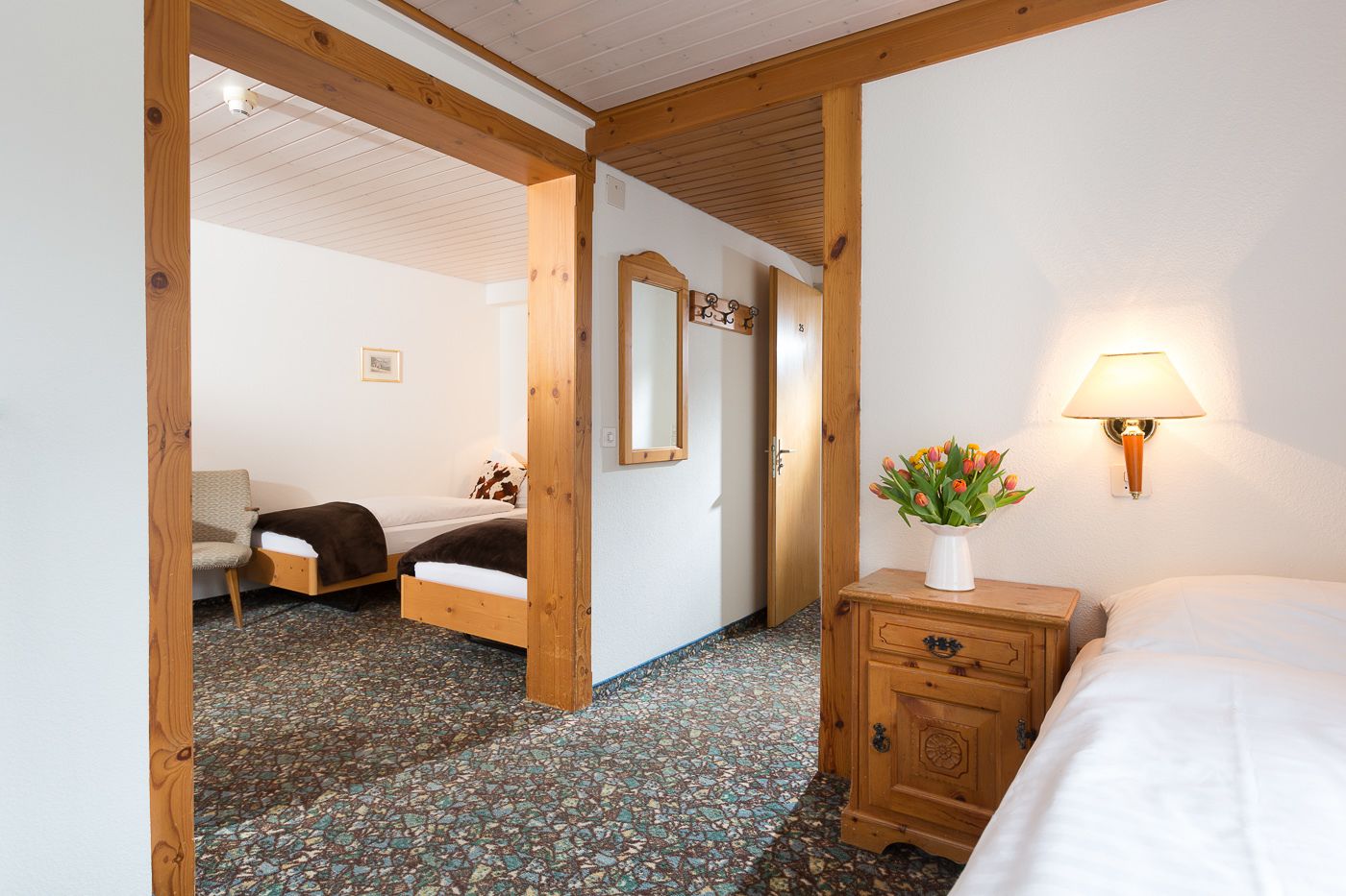 A hotel room with a bed, a chair, a wooden dresser, a mirror, and a vase with flowers.