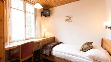 A small, cozy room with a bed, a desk, and a chair. There is a television mounted on the wall and a window with curtains.