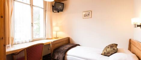 Une petite pièce agréable et confortable, avec un lit, un bureau et une chaise. Un téléviseur est fixé au mur et il y a une fenêtre avec des rideaux.