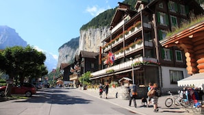 Een traditioneel hotel in chaletstijl met meerdere balkons, een berg op de achtergrond en een straat met voetgangers en geparkeerde auto's.