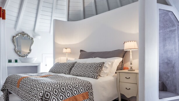 A bedroom with a sloped ceiling, a bed with patterned bedding, bedside lamps, a mirror, and a small bedside table. - Absolute Mykonos Suites & More (Mykonos)