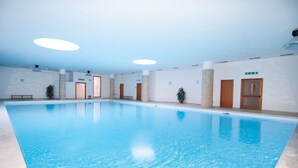 Una gran piscina interior con agua cristalina y azul, rodeada de paredes blancas y varias puertas.