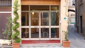 Entrada de hotel con alfombra roja, puertas de cristal y plantas en macetas a ambos lados.