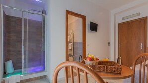 Un baño moderno con cabina de ducha de vidrio, espejo con marco de madera y una mesita con una cesta de frutas y un vaso de zumo de naranja.