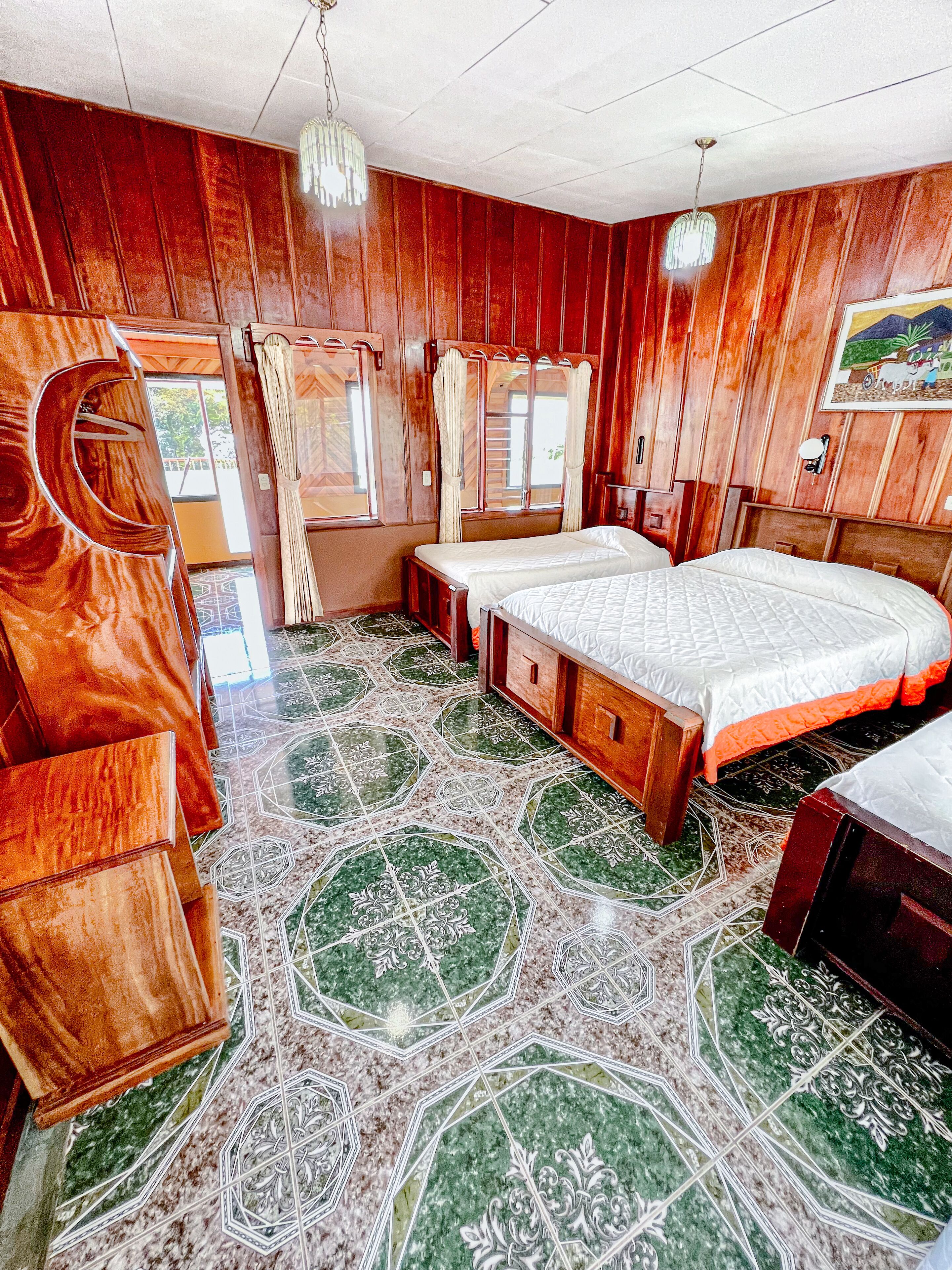 Una habitación con paredes de madera y suelo estampado. Hay dos camas con ropa blanca y un banco.