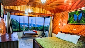 A bedroom with a bed, a bench, and a view of a tower through large windows. - Hotel Don Taco (Monteverde)