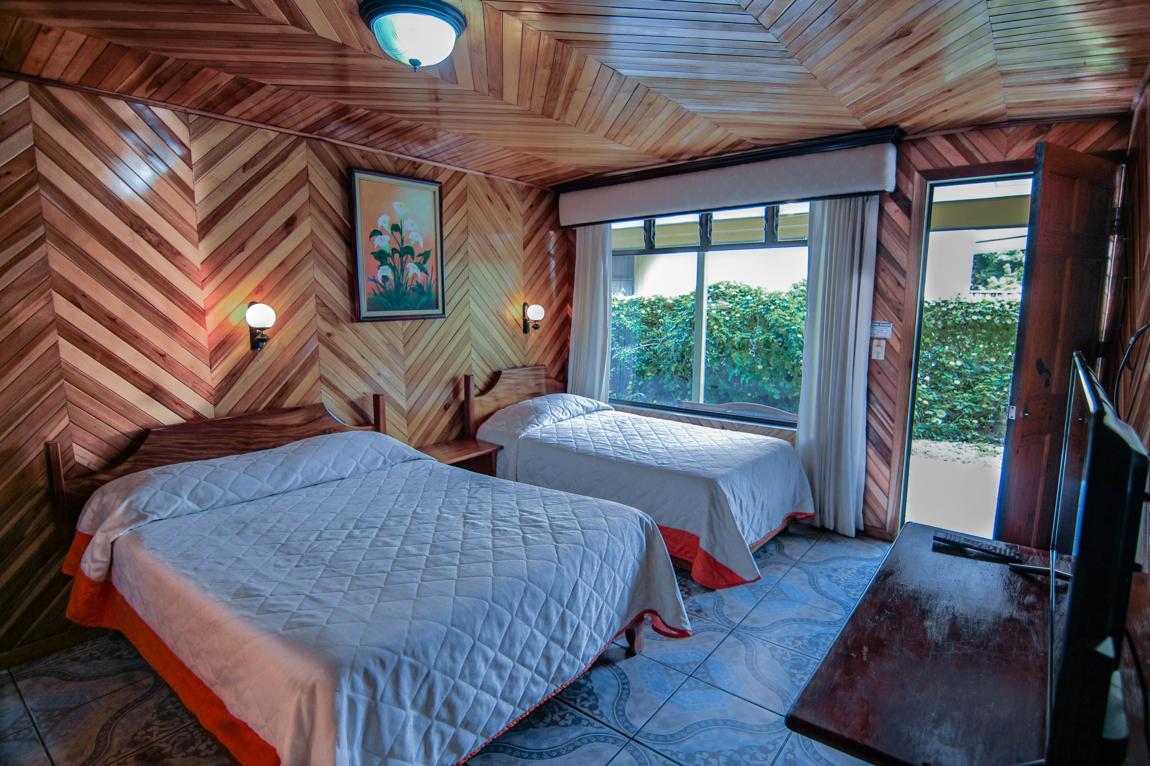 A room with two beds, wooden walls, a large window, and a view of greenery outside.