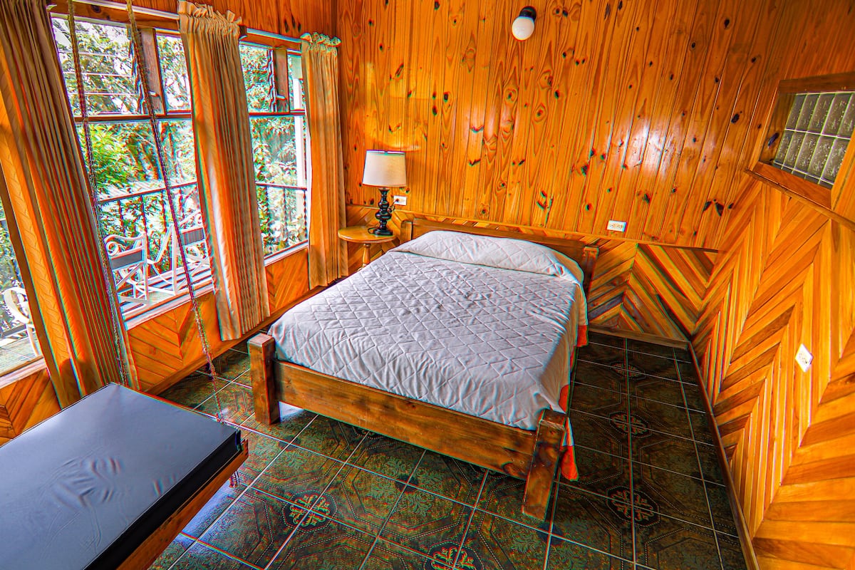 Una habitación de madera con una cama, una mesa y un ventanal con cortinas.