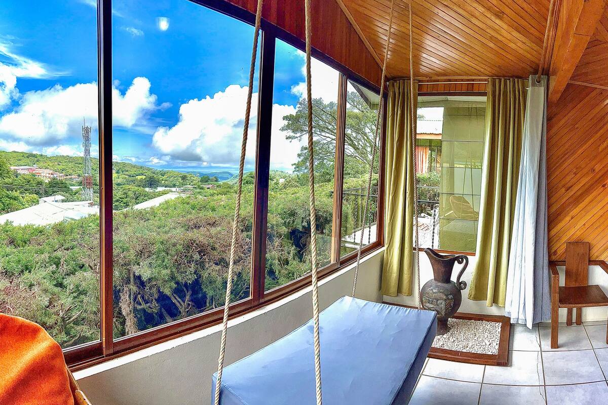 Una habitación con un ventanal grande que ofrece vistas a una zona boscosa, una cama con almohadas, una silla y una mesita.