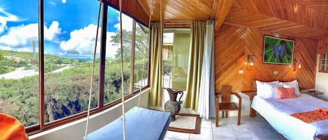 A room with a large window offering a view of a forested area, a bed with pillows, a chair, and a small table.