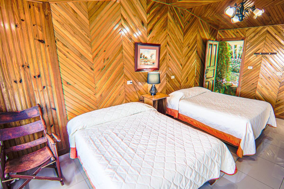 Una habitación con paredes de madera, dos camas, una mesita de noche con lámpara y una silla.