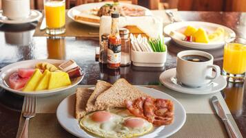 Una mesa de desayuno con platos de huevos, tostadas, tocino y fruta. También hay condimentos, una taza de café y un vaso de jugo de naranja.