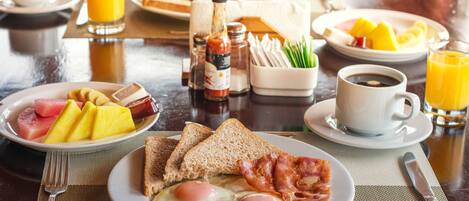 A breakfast table with plates of eggs, toast, bacon, and fruit. There are also condiments, a coffee cup, and a glass of orange juice.