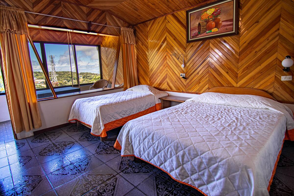 Habitación de hotel con dos camas, un ventanal con cortinas, un revestimiento de pared de madera en espiga y un cuadro sobre las camas.