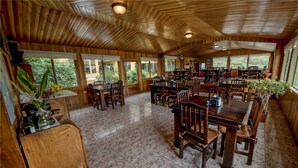 A spacious dining area with wooden tables and chairs, large windows offering views of greenery, and a high ceiling with wooden beams. - Hotel Don Taco (Monteverde)