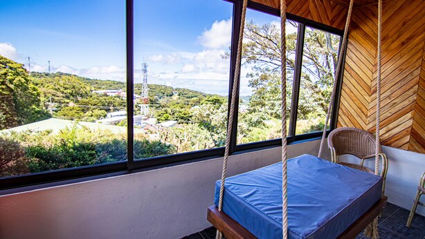 A room with a swing bed, large windows, and a view of a green landscape. - Hotel Don Taco (Monteverde)