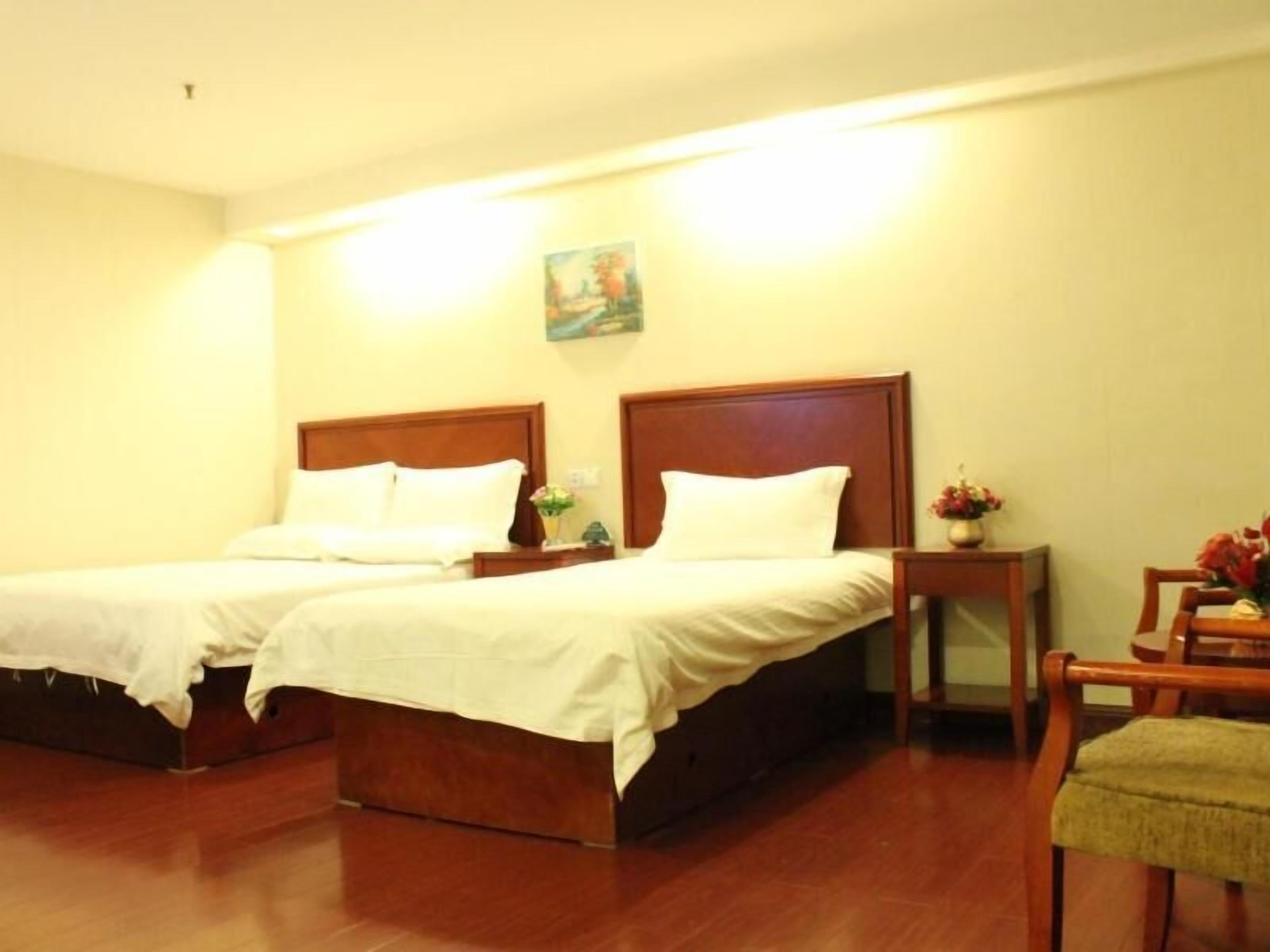 A hotel room with two beds, wooden headboards, a small table with a vase, and a chair.