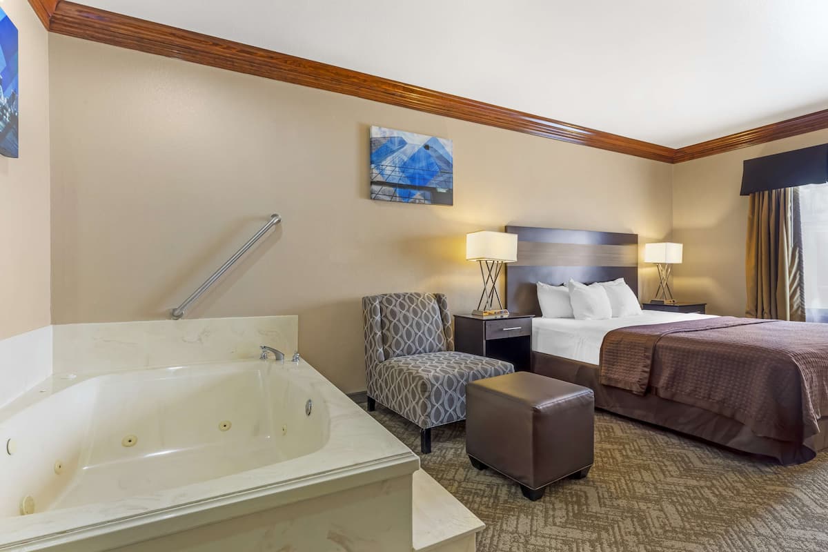 Ein Hotelzimmer mit einer großen Badewanne, einem Bett mit brauner Tagesdecke, einem grau gemusterten Sessel, zwei Nachttischlampen und einem Bild an der Wand.