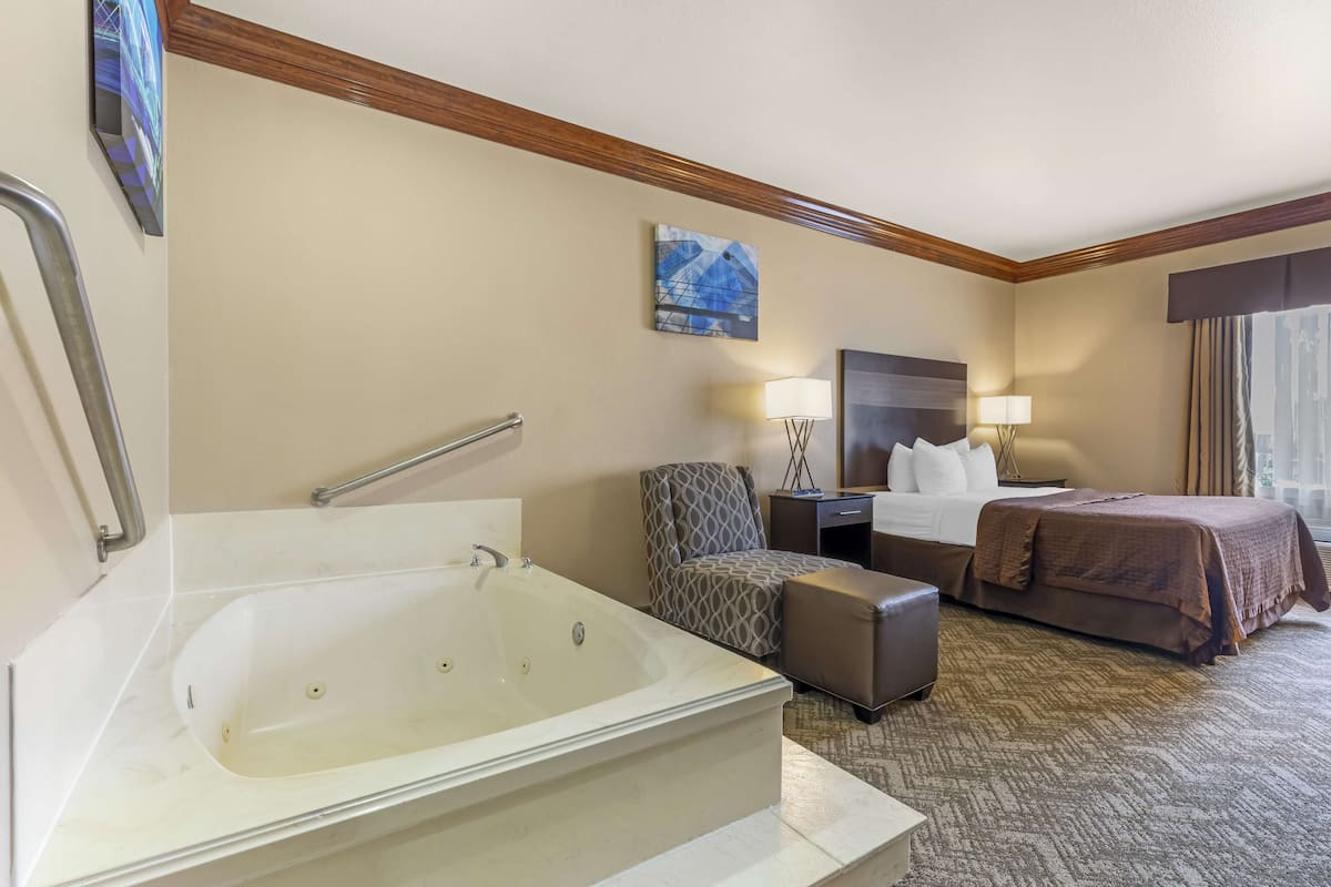 Ein Hotelzimmer mit einer großen Badewanne, einem Bett mit brauner Bettdecke, einem gemusterten Sessel und einem Nachttisch mit Lampe.