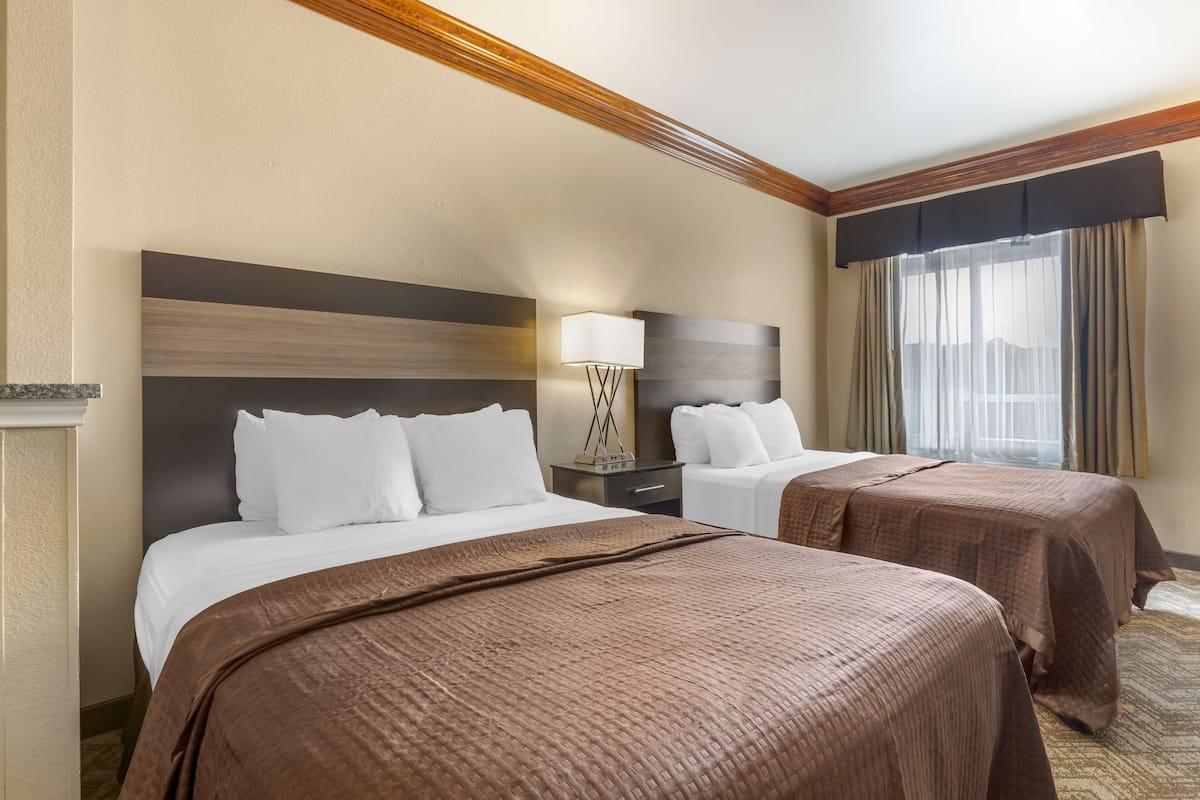A hotel room with two beds, a nightstand, a lamp, and a window with curtains.