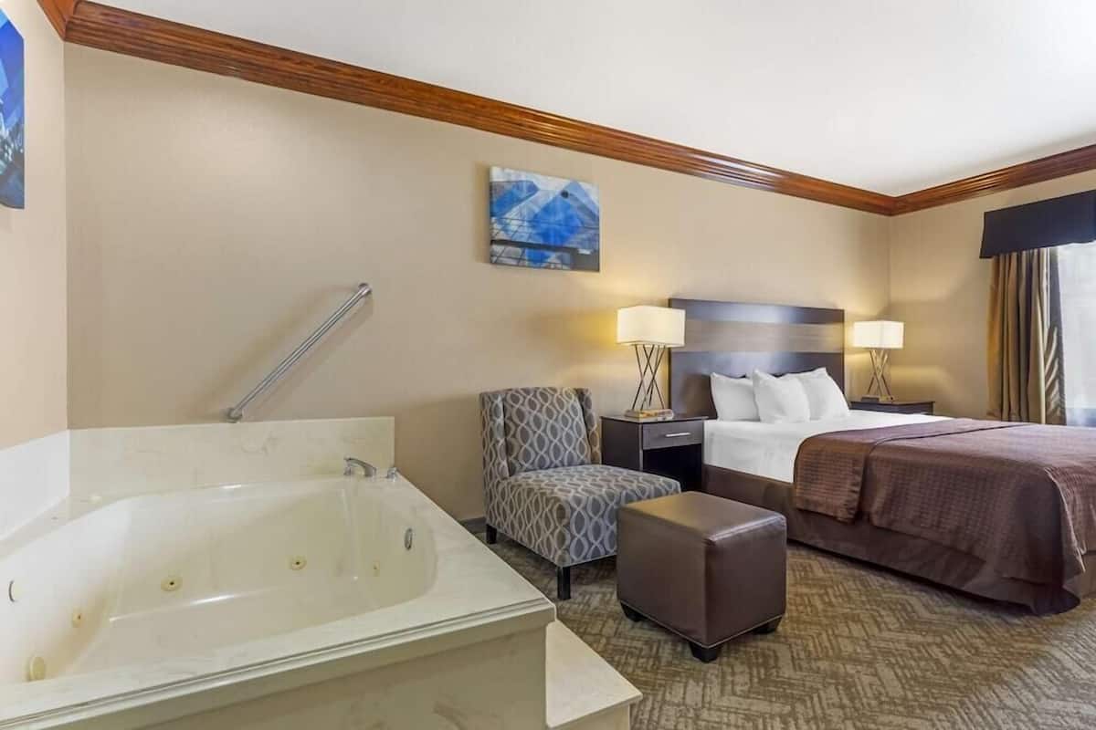 A hotel room with a large bathtub, a bed with a brown bedspread, a patterned armchair, two bedside tables with lamps, and a painting on the wall.
