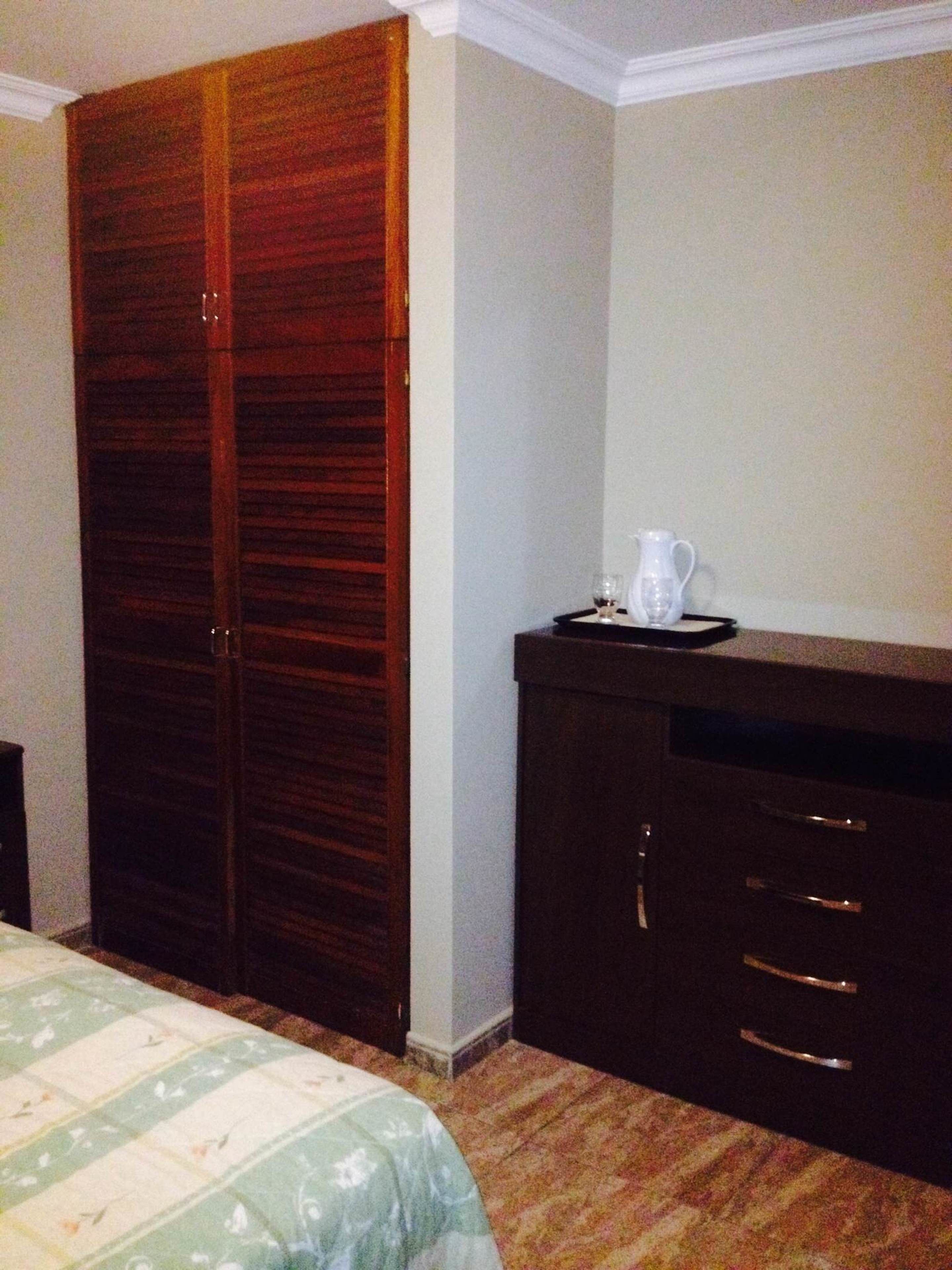 A bedroom with a large wooden wardrobe, a bed with a floral bedspread, and a small table with a white pitcher and glass.
