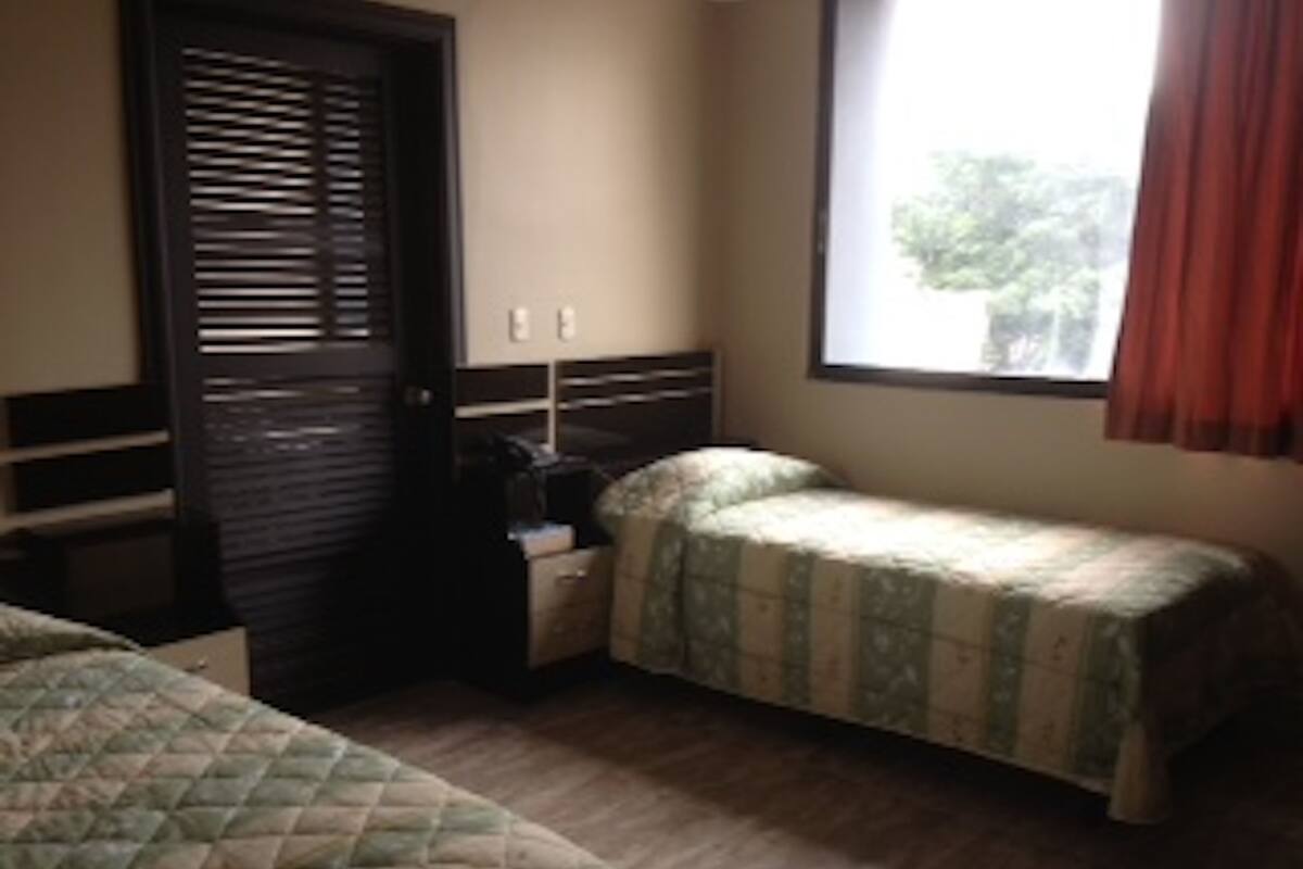 A room with two beds, a nightstand, a window with curtains, and a door with blinds.