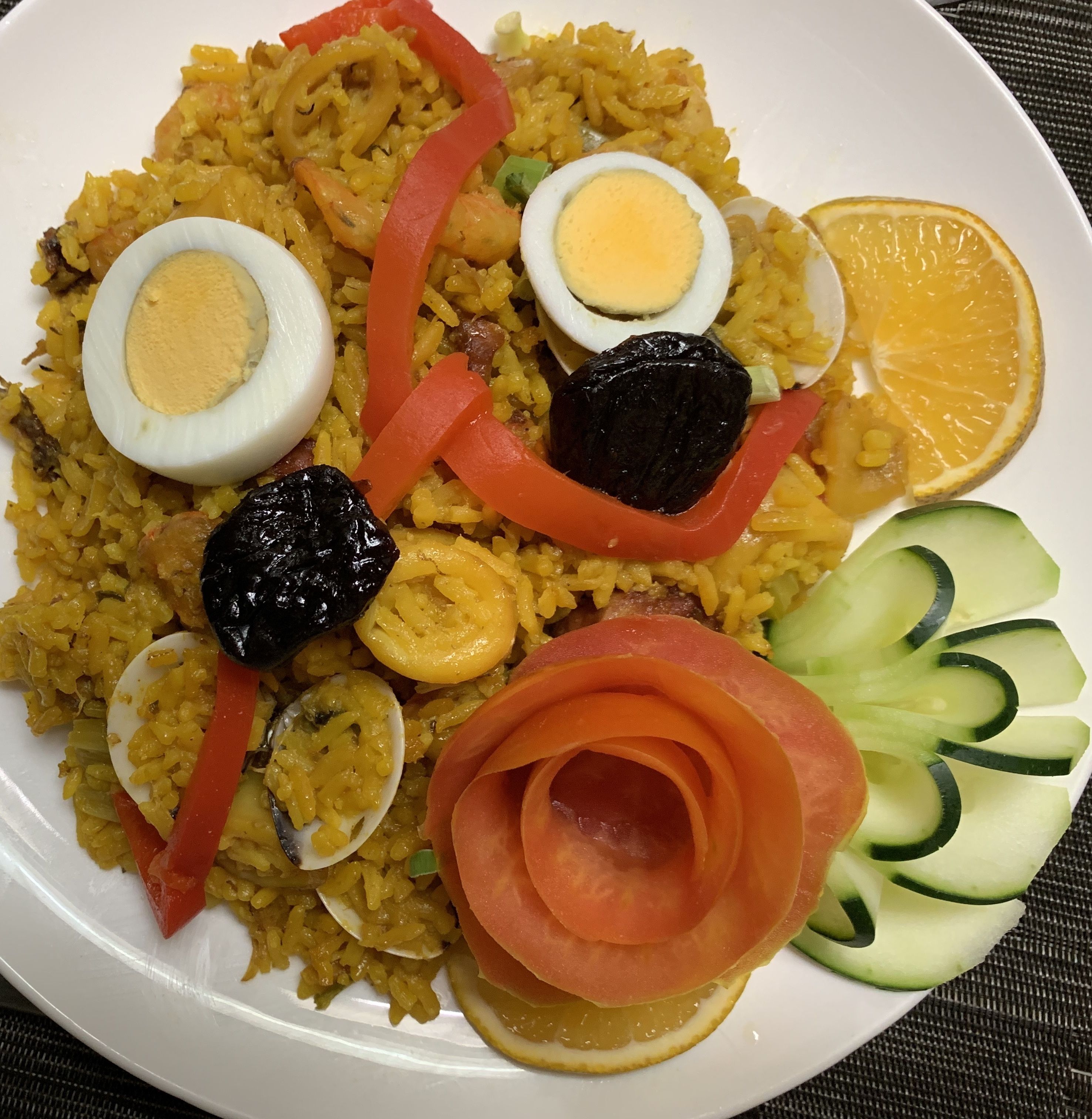 A plate of yellow rice with slices of boiled egg, red bell pepper, black olives, and cucumber slices.