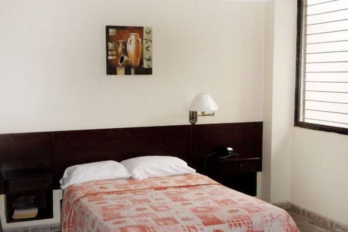 A hotel room with a bed, a nightstand, a lamp, a window with blinds, and a wall-mounted picture.