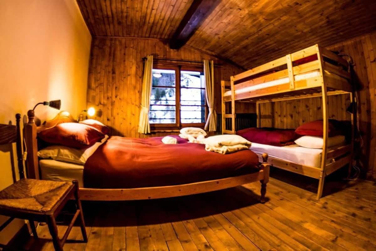 A wooden cabin room with a bunk bed, a single bed, a chair, and a window with curtains.
