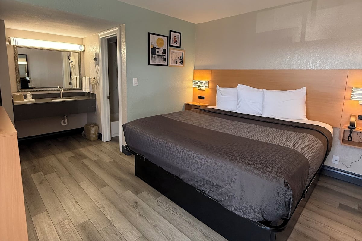 A hotel room with a large bed, a vanity with a mirror, a wooden floor, and a wall-mounted light fixture.