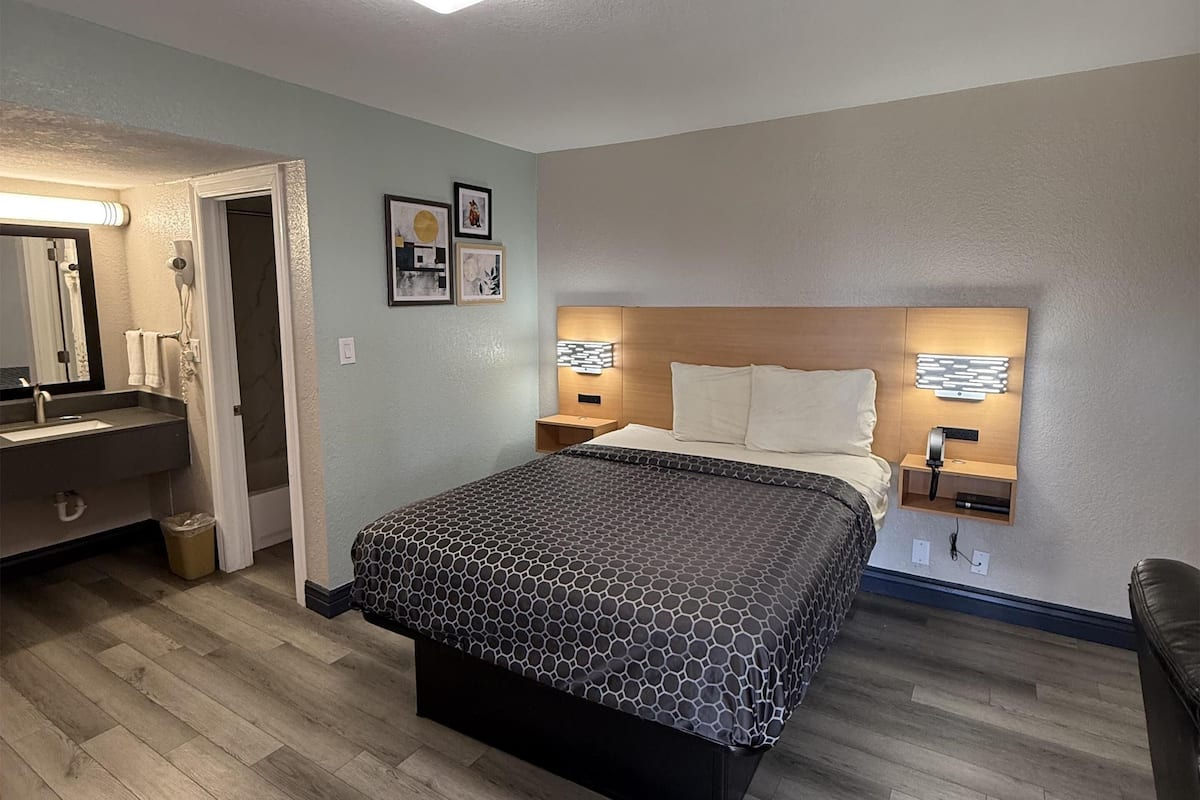 A hotel room with a bed, a bedside table, a nightstand, a mirror, a sink, and a trash bin.