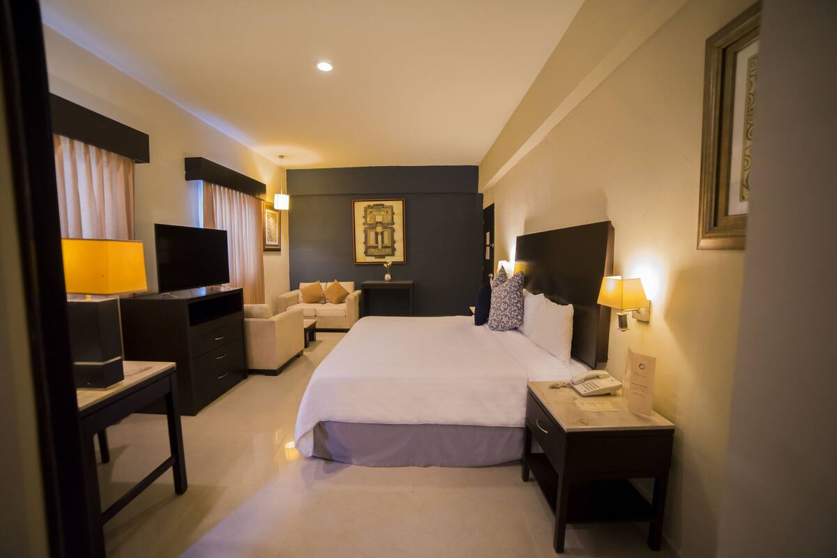 A hotel room with a large bed, a TV, a desk with a lamp, a chair, and a nightstand with a phone.