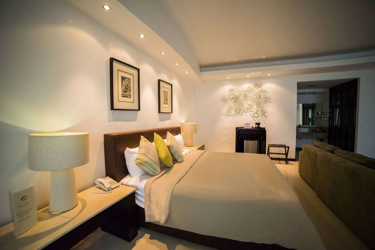 A modern hotel room with a large bed, bedside lamps, a small table with a phone, a wall-mounted plant, and a view into the kitchen.