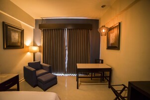 Una habitación de hotel con cama, escritorio, silla, mesa, lámpara y ventana con cortinas.