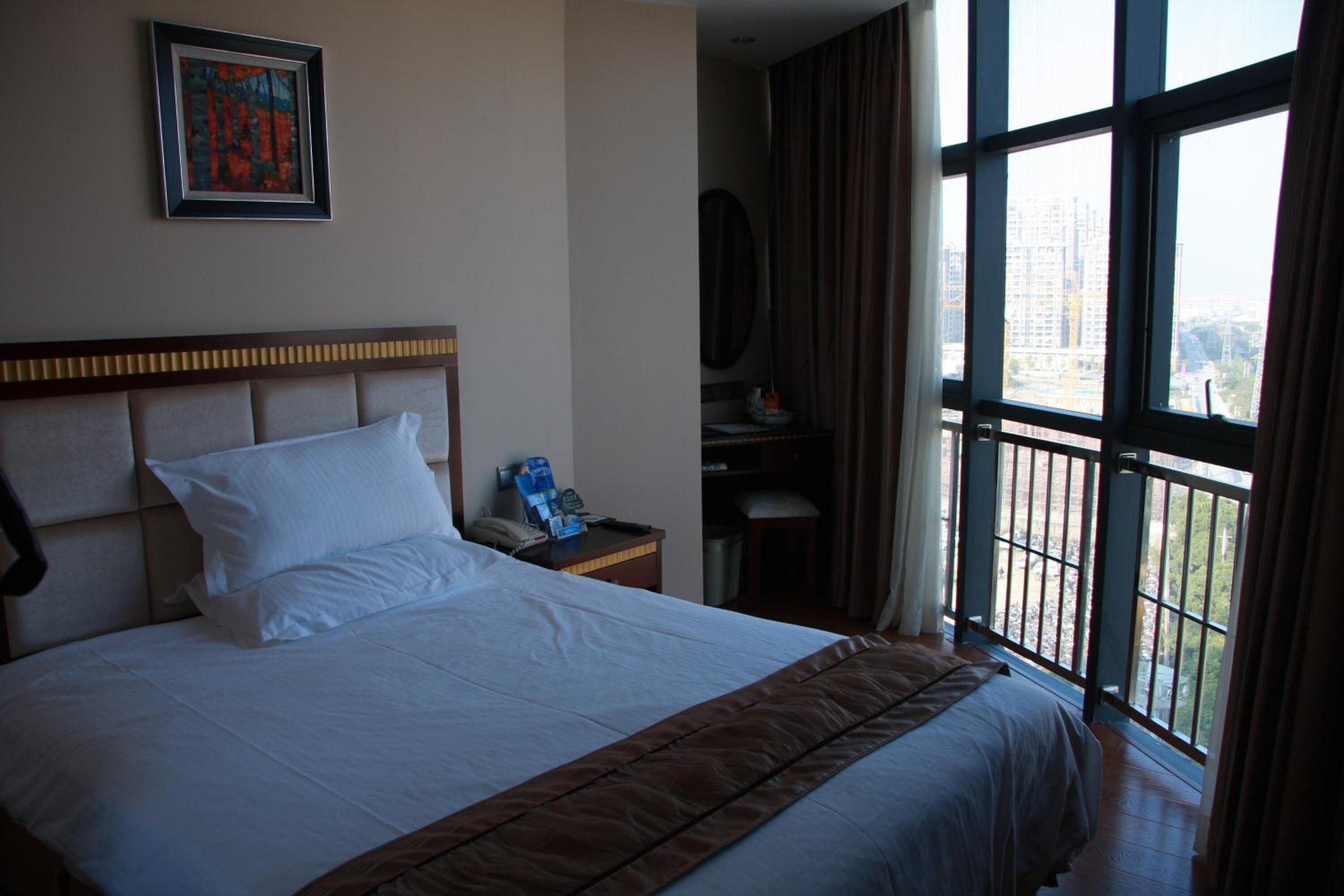 A hotel room with a large bed, a nightstand, a desk, and a window with a view of buildings.